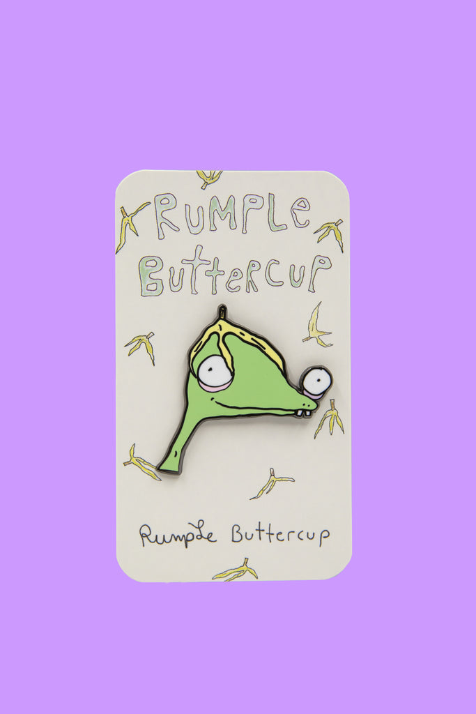Enamel pin of Rumple Buttercup's face wearing banana hat in front of paper card covered in hand-drawn bananas.