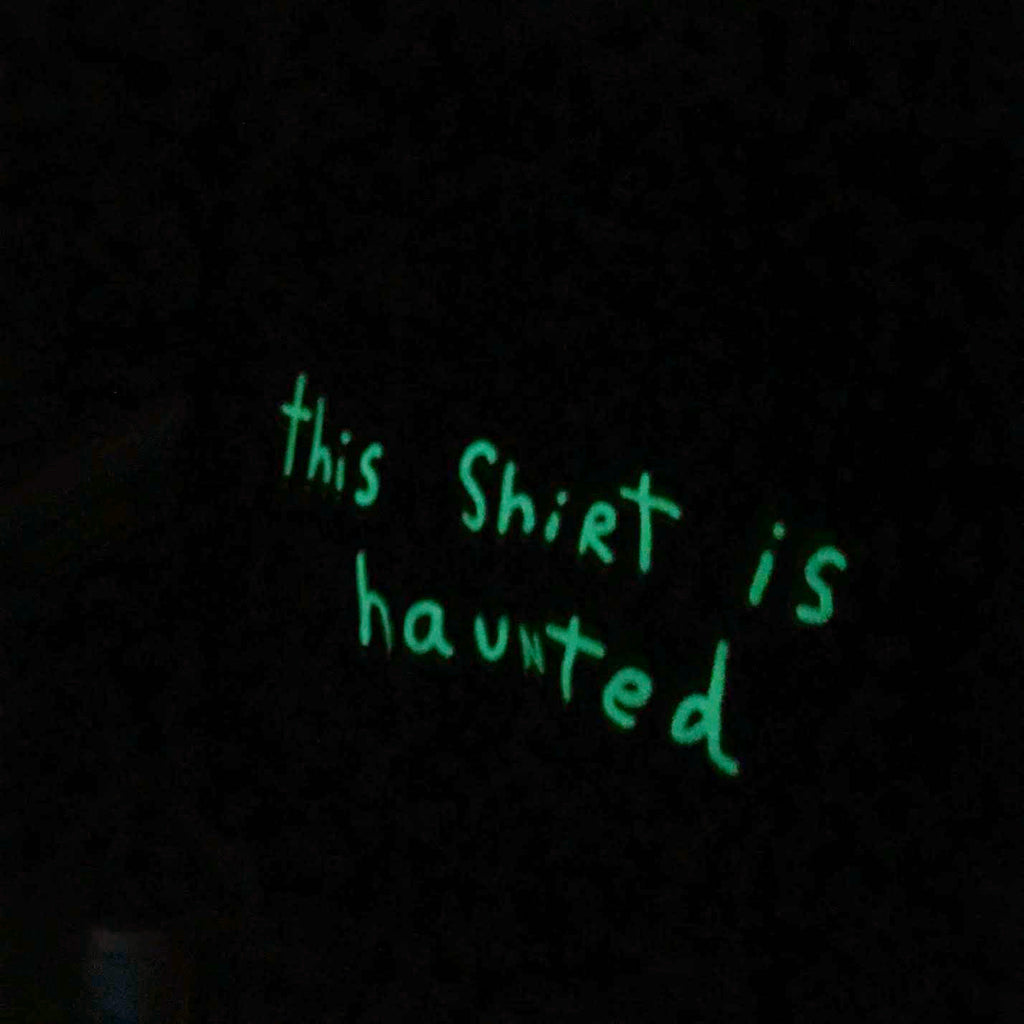Green glowing handwriting on it that says “this shirt is haunted”.