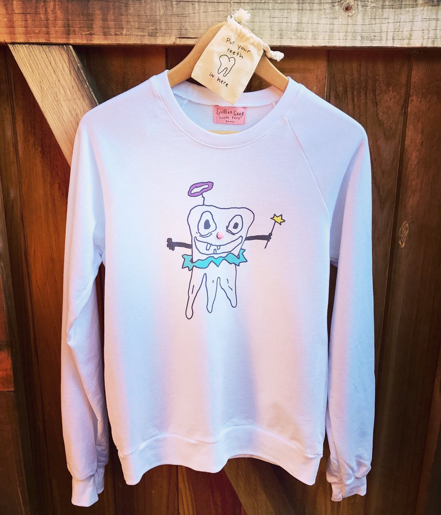 White crew neck sweatshirt on hanger with hand drawn artwork of a tooth fairy, holding a wand. Small canvas pouch above the sweatshirt with handwritten text that says “Put your teeth in here”.