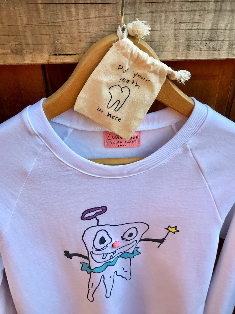 White crew neck sweatshirt on hanger with hand drawn artwork of a tooth fairy, holding a wand. Small canvas pouch above the sweatshirt with handwritten text that says “Put your teeth in here”.