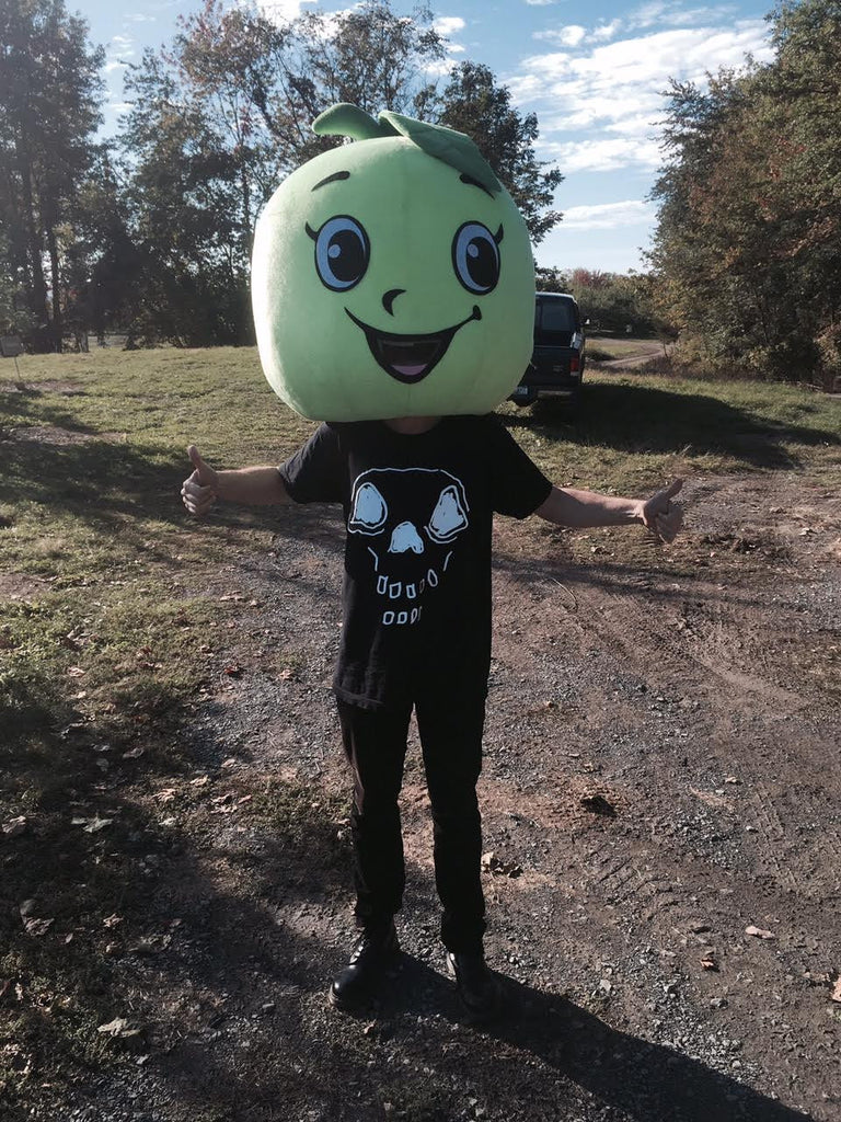 Picture of Matthew standing in nature, wearing a hand-drawn skeleton head shirt while wearing a giant apple on his head.