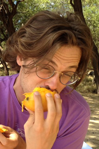 Picture of Matthew with glasses, eating fruit under a tree while wearing a purple shirt. 